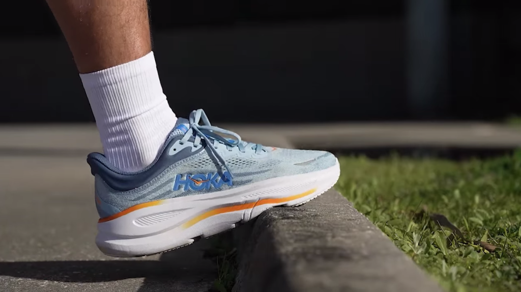 Sepatu lari HOKA digunakan pelari saat berdiri di tepi trotoar dengan desain midsole tebal dan ringan.