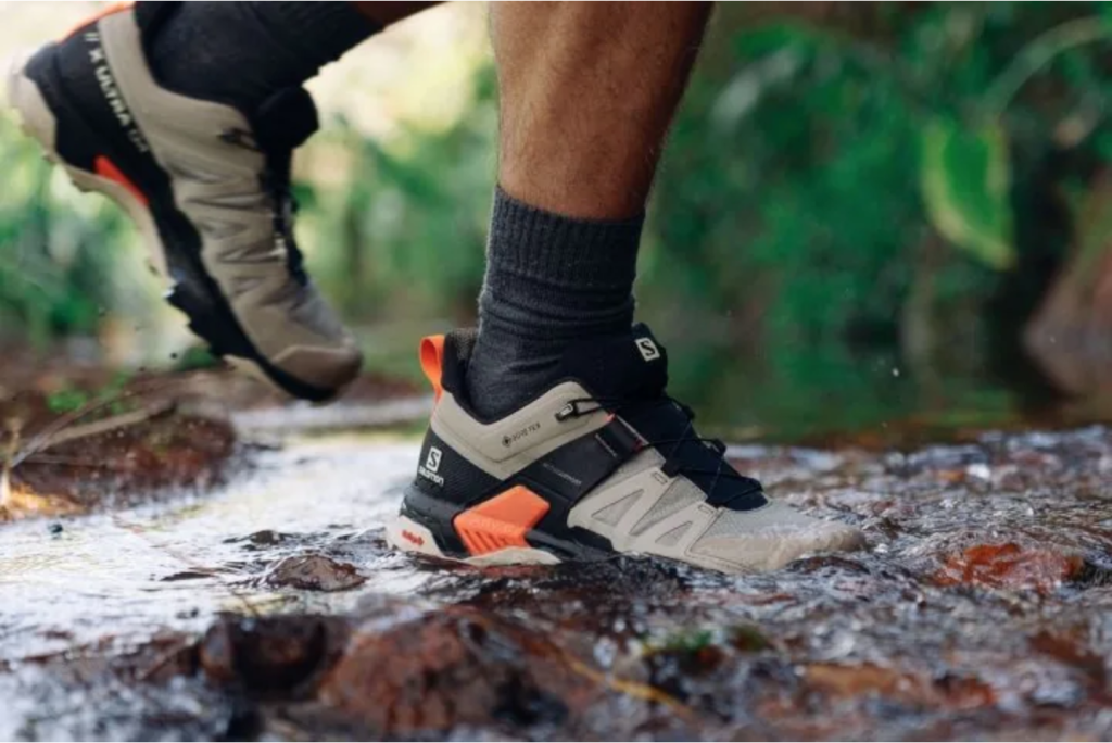 Sepatu hiking Salomon digunakan menyeberangi aliran air dangkal untuk menunjukkan kemampuan waterproof dan grip.