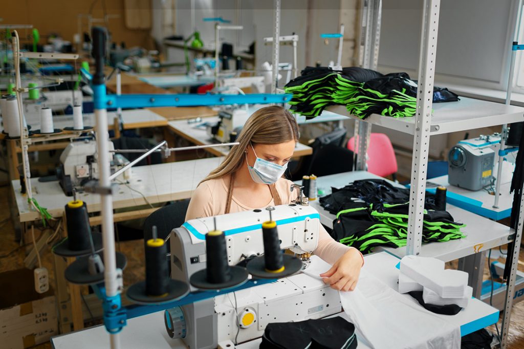 Young woman working as a tailor in a sewing workshop, wearing a mask and sewing garments.